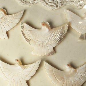 May include: A collection of carved, off-white bird figurines with outstretched wings. Each bird has a small, red accent on its head. The wings are detailed with etched lines, and the design is delicate and intricate. The figurines are likely made of shell.