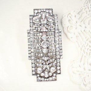 May include: A silver-toned, geometric brooch with a detailed design featuring clear rhinestones. The brooch has a rectangular shape with a central floral motif.