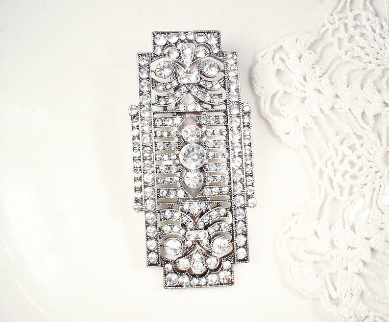 May include: A silver-toned, geometric brooch with a detailed design featuring clear rhinestones. The brooch has a rectangular shape with a central floral motif.