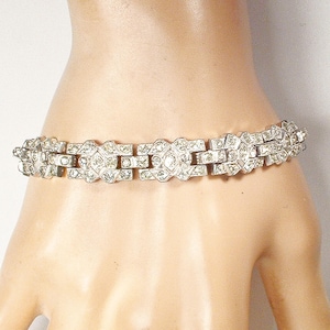 May include: A silver-toned bracelet with a geometric design, featuring multiple rectangular and diamond-shaped links. The bracelet is adorned with small, clear gemstones, creating a sparkling effect. The jewelry is displayed on a light skin tone.