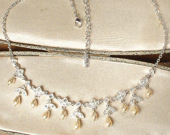 Peut inclure: Un collier argenté au design délicat. Il présente des accents clairs et étincelants et des perles en forme de goutte d'eau de couleur crème. Le collier est présenté sur une surface de couleur claire.