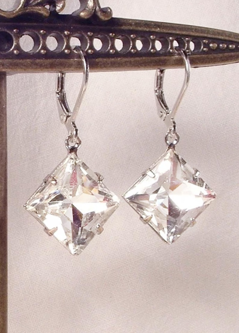 Puede incluir: Un par de pendientes cuadrados de cristal transparente con cierres de palanca plateados. Los pendientes se exhiben en un soporte met&aacute;lico decorativo, mostrando sus superficies facetadas y su brillo. Los pendientes est&aacute;n dise&ntilde;ados para ser usados.