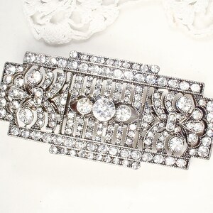 May include: A silver-toned brooch with a geometric design, featuring a central cluster of clear rhinestones and smaller rhinestones throughout.