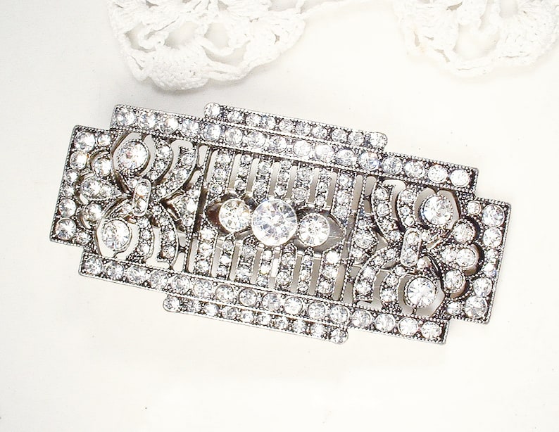 May include: A silver-toned brooch with a geometric design, featuring a central cluster of clear rhinestones and smaller rhinestones throughout.