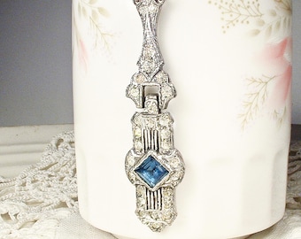 May include: A silver-toned pendant necklace with an Art Deco design. The pendant features clear crystals and a square, blue gemstone. The necklace is hanging in front of a white mug with a floral design.