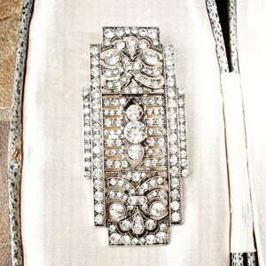 May include: An Art Deco style brooch in silver and crystal, featuring a geometric design. The brooch is set with clear rhinestones and has a delicate, intricate pattern.