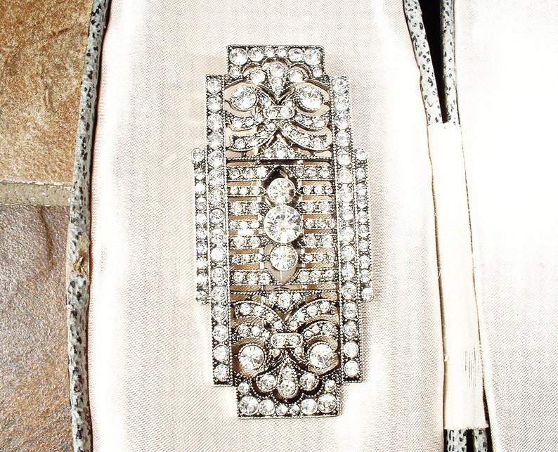 May include: An Art Deco style brooch in silver and crystal, featuring a geometric design. The brooch is set with clear rhinestones and has a delicate, intricate pattern.