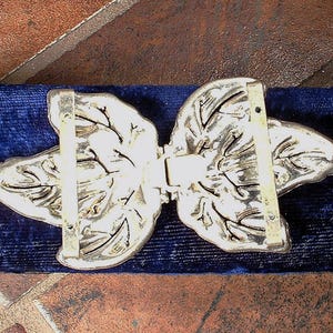 Peut inclure: Boucle de ceinture en m&eacute;tal argent&eacute; avec un motif de feuilles. La boucle est compos&eacute;e de deux moiti&eacute;s sym&eacute;triques qui se rejoignent au centre. Les feuilles pr&eacute;sentent des nervures d&eacute;taill&eacute;es et sont pos&eacute;es sur un fond de velours bleu fonc&eacute;.