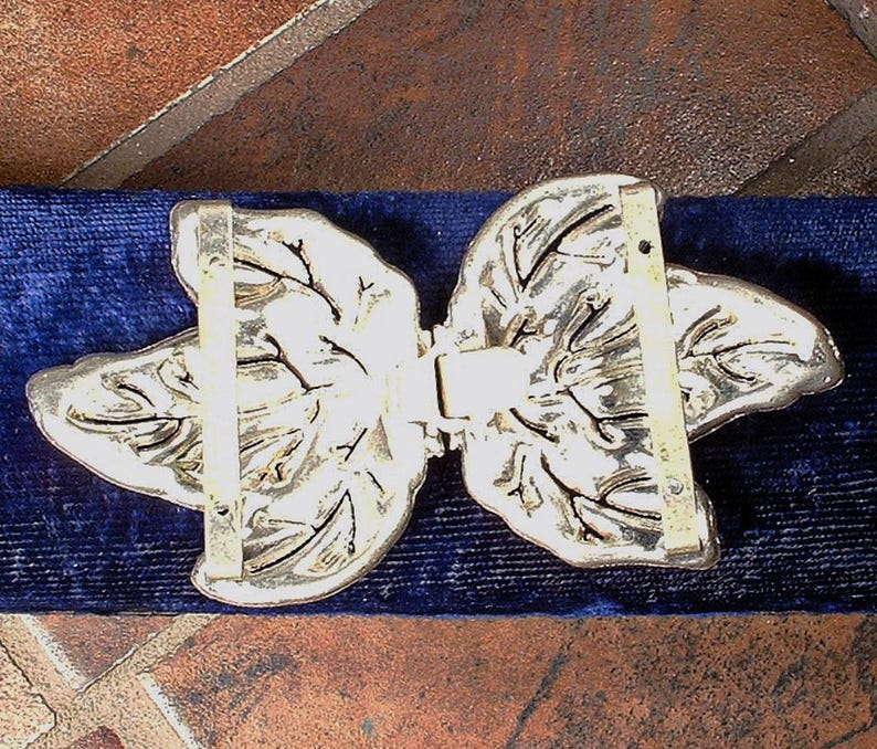 Peut inclure: Boucle de ceinture en m&eacute;tal argent&eacute; avec un motif de feuilles. La boucle est compos&eacute;e de deux moiti&eacute;s sym&eacute;triques qui se rejoignent au centre. Les feuilles pr&eacute;sentent des nervures d&eacute;taill&eacute;es et sont pos&eacute;es sur un fond de velours bleu fonc&eacute;.