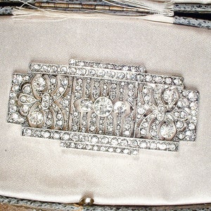 May include: A silver-toned brooch with a geometric design, featuring a central motif of a stylized flower surrounded by rows of sparkling rhinestones.