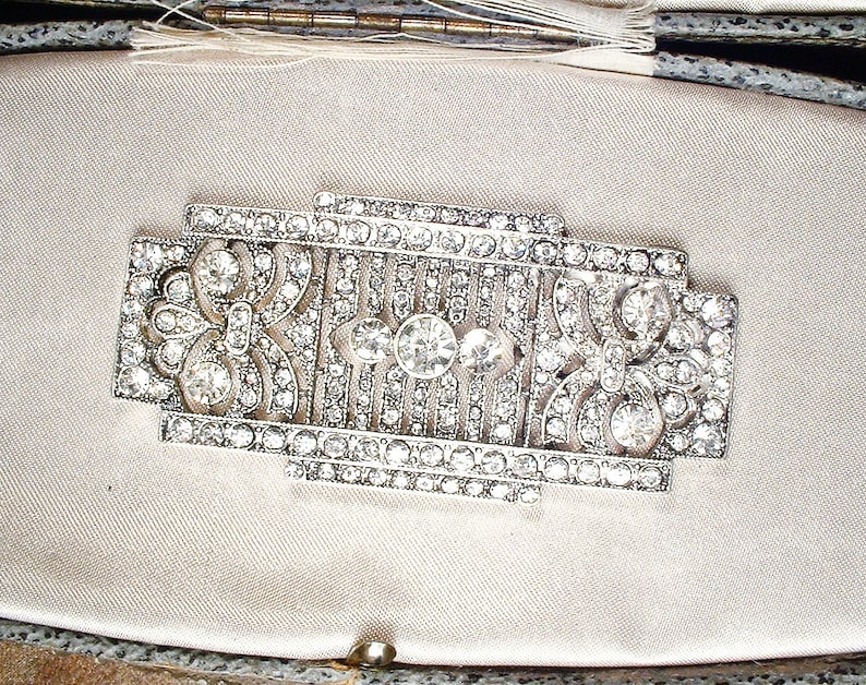 May include: A silver-toned brooch with a geometric design, featuring a central motif of a stylized flower surrounded by rows of sparkling rhinestones.