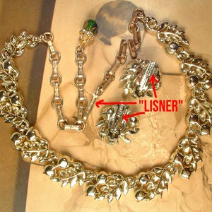 May include: A vintage gold-toned necklace and earring set. The necklace features a leaf and berry design. The earrings are clip-on style. The word "LISNER" is visible on the back of the earrings. The set is displayed on a neutral background.