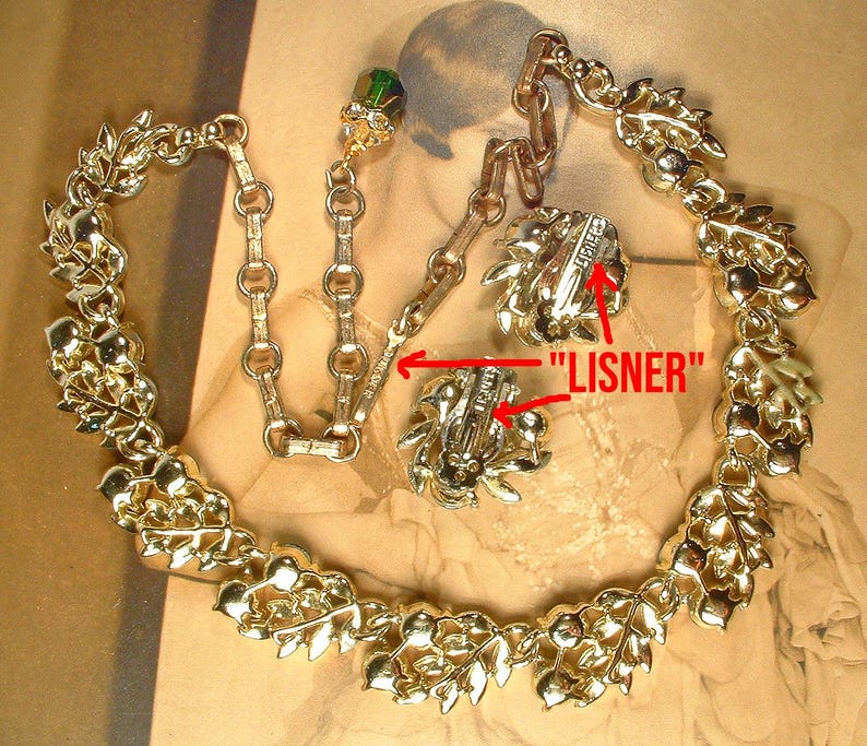 May include: A vintage gold-toned necklace and earring set. The necklace features a leaf and berry design. The earrings are clip-on style. The word "LISNER" is visible on the back of the earrings. The set is displayed on a neutral background.