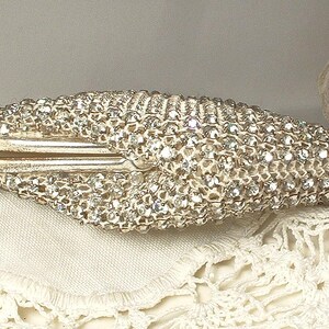 May include: A hard-shell clutch bag covered in numerous clear, reflective crystals. The exterior features a dense, textured surface of sparkling stones with a metallic frame closure visible along the center. The shape is elongated and tapered at both ends.