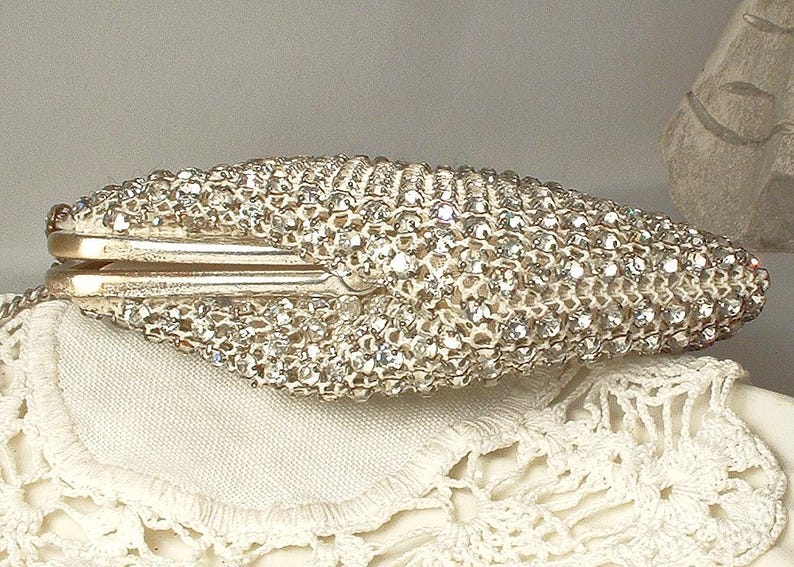 May include: A hard-shell clutch bag covered in numerous clear, reflective crystals. The exterior features a dense, textured surface of sparkling stones with a metallic frame closure visible along the center. The shape is elongated and tapered at both ends.