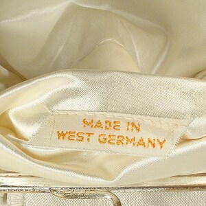 May include: The interior of a cream-colored handbag featuring a smooth satin lining. A small white fabric tag is sewn into the lining with the text MADE IN WEST GERMANY printed in gold lettering. The bag has a metallic frame visible at the bottom edge.