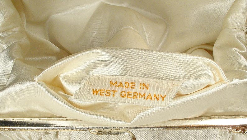 May include: The interior of a cream-colored handbag featuring a smooth satin lining. A small white fabric tag is sewn into the lining with the text MADE IN WEST GERMANY printed in gold lettering. The bag has a metallic frame visible at the bottom edge.