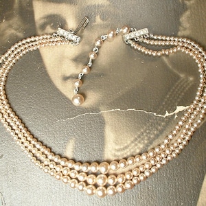 Könnte beinhalten: Eine dreireihige Perlenkette im Vintage-Stil. Die Kette besteht aus hellrosa Perlen, deren Größe variiert. Der Verschluss ist silberfarben und mit kleinen, klaren Kristallen verziert. Die Kette wird vor einem Vintage-Fotohintergrund präsentiert.