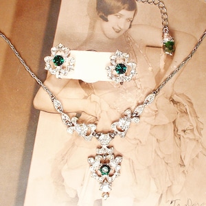 May include: A silver-toned necklace and earring set featuring intricate filigree metalwork and clear rhinestones. The necklace has a central pendant with a round emerald green stone, and the matching earrings feature the same green stone.