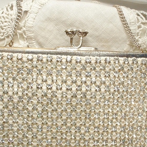 May include: A rectangular evening clutch bag covered in rows of clear, faceted rhinestones set in a silver-tone metal mesh. The bag features a silver-tone metal frame, a kiss-lock clasp closure, and a thin silver-tone metal chain shoulder strap.
