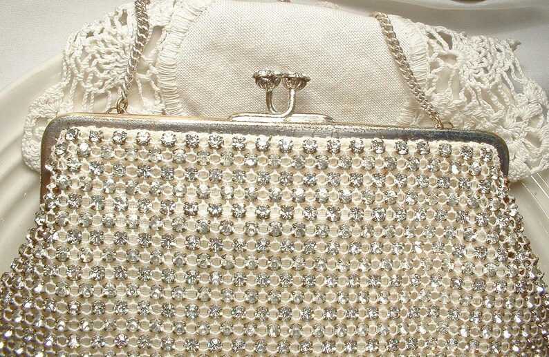 May include: A rectangular evening clutch bag covered in rows of clear, faceted rhinestones set in a silver-tone metal mesh. The bag features a silver-tone metal frame, a kiss-lock clasp closure, and a thin silver-tone metal chain shoulder strap.
