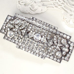 May include: A silver-toned brooch with a geometric design, featuring a central cluster of clear rhinestones and smaller rhinestones throughout.