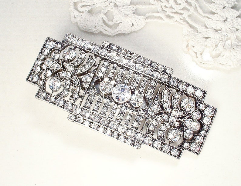 May include: A silver-toned brooch with a geometric design, featuring a central cluster of clear rhinestones and smaller rhinestones throughout.