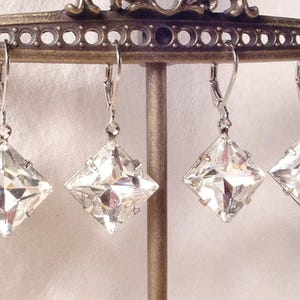 Puede incluir: Cuatro pendientes de cristal transparente, tallados en forma cuadrada, con cierres de palanca plateados, cuelgan de un expositor decorativo. Los pendientes reflejan la luz, mostrando sus superficies facetadas. Fondo blanco roto.