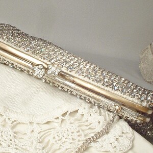 May include: A rectangular clutch bag covered in clear, sparkling rhinestones with a gold-toned metal frame and a matching gold-toned metal chain strap. The closure features two small, circular, rhinestone-encrusted accents on the top edge.
