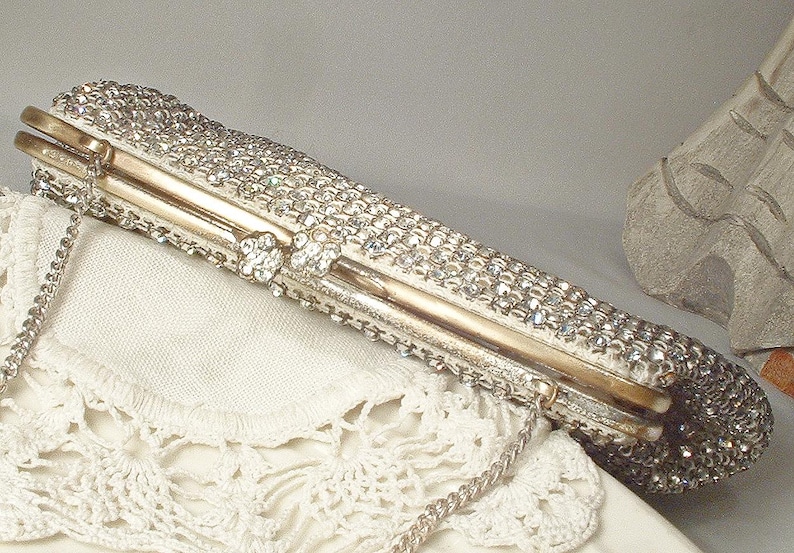 May include: A rectangular clutch bag covered in clear, sparkling rhinestones with a gold-toned metal frame and a matching gold-toned metal chain strap. The closure features two small, circular, rhinestone-encrusted accents on the top edge.