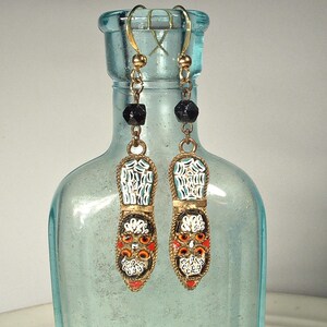 May include: A pair of mosaic earrings with a gold-tone metal frame. The earrings feature a shoe-like design with white, orange, and red mosaic details. Black beads and gold-tone hooks complete the design. The earrings hang in front of a light blue glass bottle.