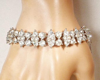 May include: A silver-toned bracelet with multiple rows of clear, sparkling rhinestones. The bracelet is designed with a series of clustered, round and oval-shaped stones, creating a dazzling effect. The bracelet is shown on a person's wrist.
