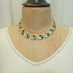 May include: A gold-toned necklace with green and blue gemstones and white leaf accents. The necklace is displayed on a mannequin wearing a white top with a delicate trim around the neckline. The necklace has a classic, elegant design.