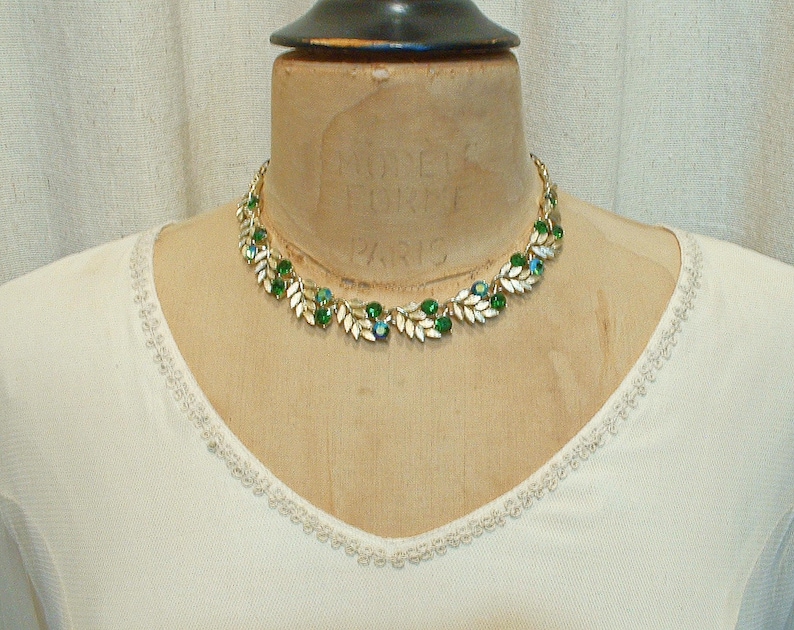 May include: A gold-toned necklace with green and blue gemstones and white leaf accents. The necklace is displayed on a mannequin wearing a white top with a delicate trim around the neckline. The necklace has a classic, elegant design.