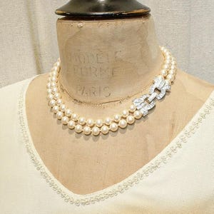 Può includere: Una collana a doppio filo con perle color crema e una chiusura centrale rettangolare color argento incastonata con pietre trasparenti scintillanti. La chiusura collega le due file di perle, creando un gioiello strutturato e simmetrico.
