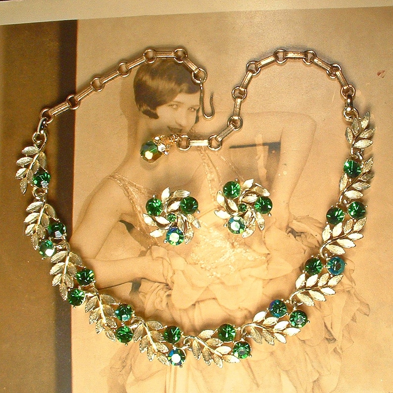 May include: A vintage jewelry set featuring a necklace and matching earrings. The necklace and earrings have a gold-tone metal leaf design, adorned with green and blue-green gemstones. The necklace is connected with a chain and clasp.