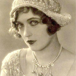 May include: A vintage-style cloche hat covered in intricate white beadwork, paired with a matching beaded garment and a multi-strand pearl necklace featuring dangling pearl accents. The text Amore Treasure is written in black script across the bottom.