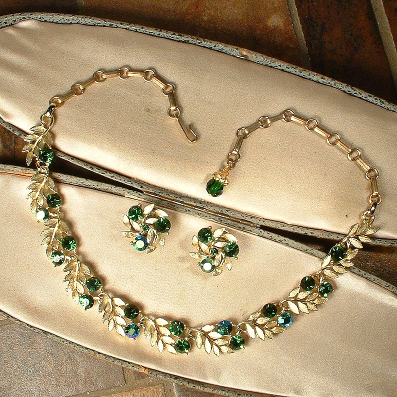 May include: A vintage jewelry set featuring a gold-toned necklace and matching earrings. The necklace and earrings are adorned with green and blue gemstones and leaf-shaped accents. The set is displayed on a satin-lined surface.