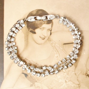 Puede incluir: Un brazalete plateado vintage con tres filas de brillantes pedrería transparente. El brazalete se encuentra sobre una fotografía en tonos sepia de una mujer, creando una estética clásica y elegante. El cierre también está adornado con pedrería.