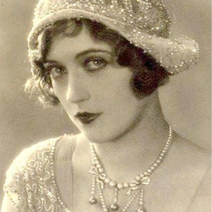 May include: A black and white vintage photograph of a woman wearing a beaded hat and a pearl necklace. The woman is looking at the camera with a serious expression. The text "Amore Treasure" is written in cursive at the bottom of the image.