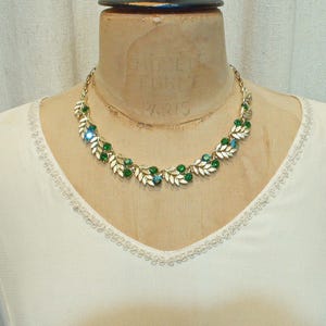 May include: A gold-toned necklace with a leaf and berry design. The necklace features green and blue gemstones, and is displayed on a mannequin. The necklace is a vintage piece.