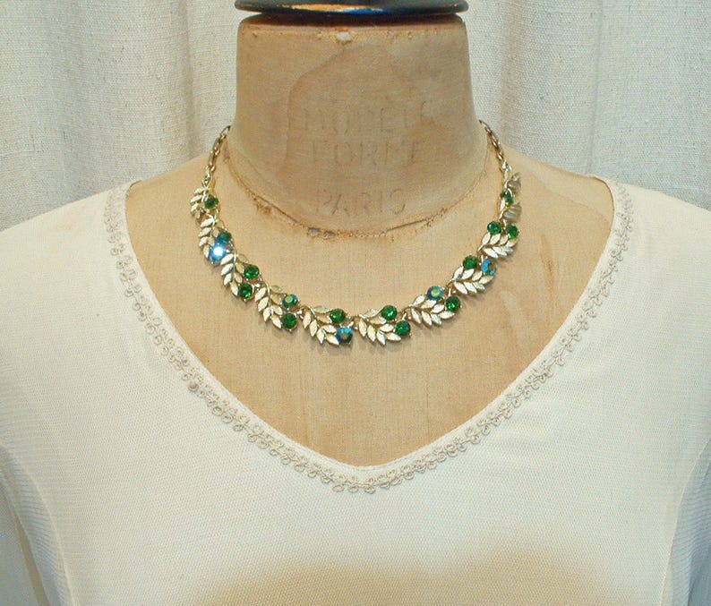 May include: A gold-toned necklace with a leaf and berry design. The necklace features green and blue gemstones, and is displayed on a mannequin. The necklace is a vintage piece.