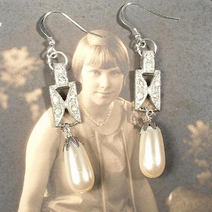 May include: A pair of silver dangle earrings with a rectangular design featuring clear rhinestones and a pearlescent drop.