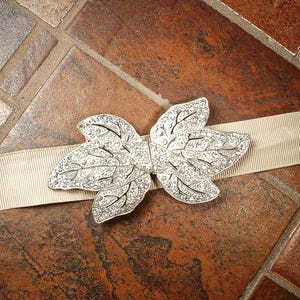 Peut inclure: Un ornement argent&eacute; en forme de feuille avec des d&eacute;tails complexes, pos&eacute; sur un ruban beige. La pi&egrave;ce d&eacute;corative pr&eacute;sente une surface scintillante et textur&eacute;e, id&eacute;ale pour la mode ou l'artisanat.
