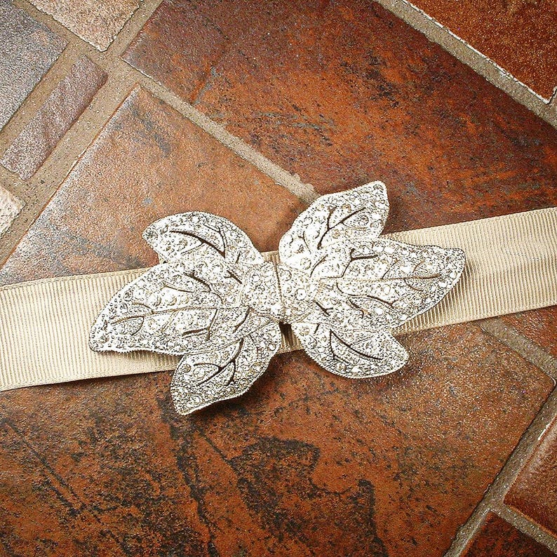 Peut inclure: Un ornement argent&eacute; en forme de feuille avec des d&eacute;tails complexes, pos&eacute; sur un ruban beige. La pi&egrave;ce d&eacute;corative pr&eacute;sente une surface scintillante et textur&eacute;e, id&eacute;ale pour la mode ou l'artisanat.