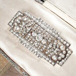 May include: A silver and crystal brooch with a geometric design. The brooch is set with clear rhinestones and has a delicate, intricate pattern.