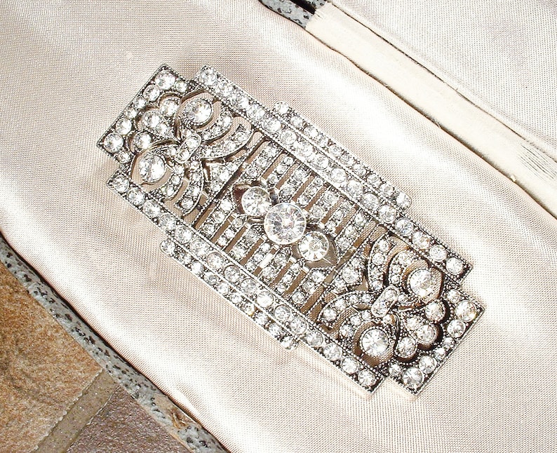 May include: A silver and crystal brooch with a geometric design. The brooch is set with clear rhinestones and has a delicate, intricate pattern.