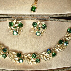May include: A vintage jewelry set featuring a gold-toned necklace, earrings, and pendant. The necklace and earrings have a leaf design with emerald green gemstones. The pendant has a single green gemstone. The set is displayed in a beige jewelry box.