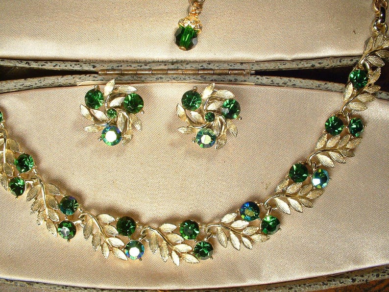 May include: A vintage jewelry set featuring a gold-toned necklace, earrings, and pendant. The necklace and earrings have a leaf design with emerald green gemstones. The pendant has a single green gemstone. The set is displayed in a beige jewelry box.