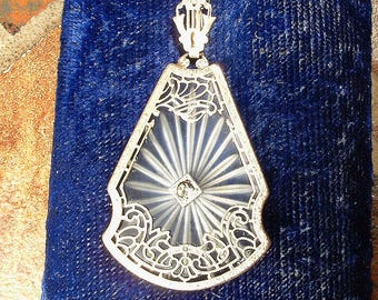 May include: A silver-toned pendant necklace featuring a translucent, fan-shaped glass insert with a sunburst pattern radiating from a central clear stone. The glass is encased in an intricate, openwork metal frame with decorative scrollwork.